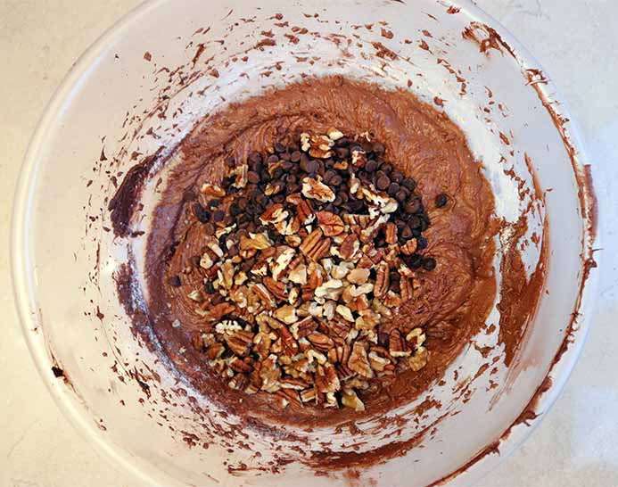 A mixing bowl filled with cake batter and chocolate chips and pecan nuts and walnuts