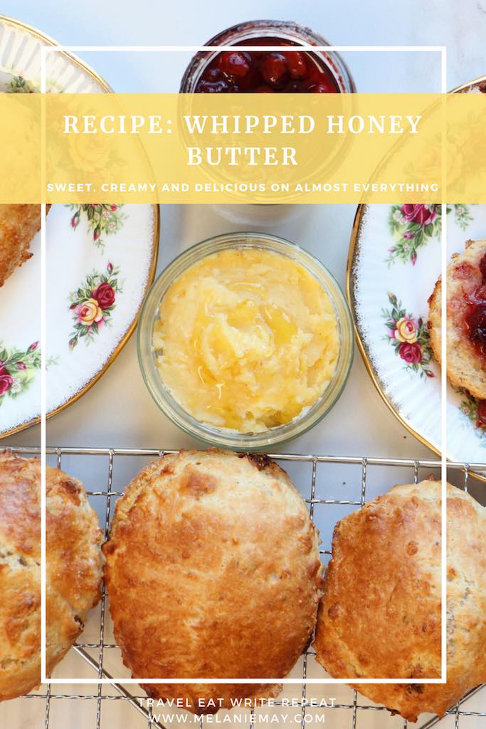 A pot of honey butter with honey drizzled on top beside some homemade scones and jam.