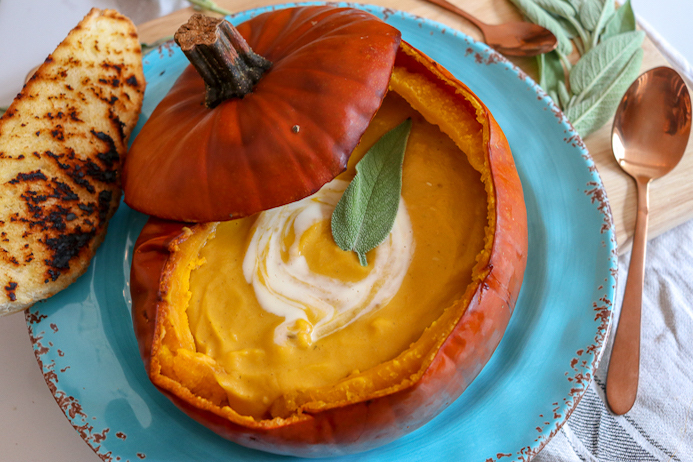 Pumpkin Soup Recipe served in a pumpkin shell garnished with cheese, cream and herbs.